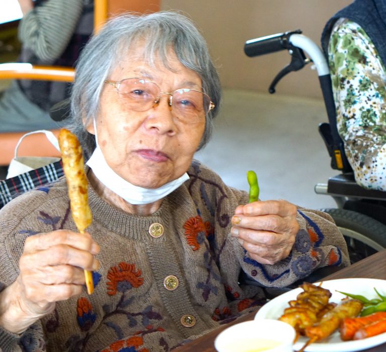 高齢者が食事イベントで串焼きを持ち、笑顔で参加している様子